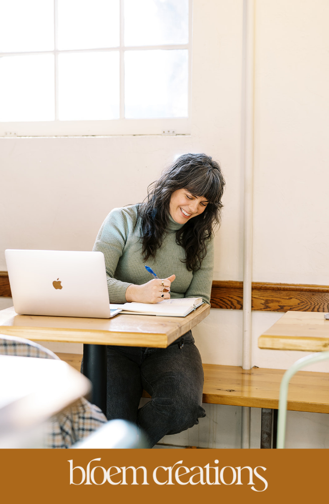 Carrie of Bloem Creations sits at a table with her laptop and writing in a notepad.